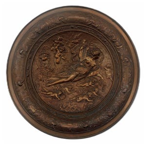 May include: A round, bronze-colored decorative plate with a detailed relief sculpture. The central image depicts a woman playing a harp, surrounded by cherubs and floral elements. The outer rim features an ornate, repeating pattern.
