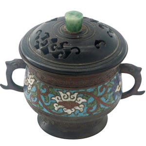 May include: An antique bronze incense burner with a lid featuring intricate cut-out designs. The body is adorned with colorful floral patterns in blue, red, and white. It has two handles and a jade stone on top. A decorative piece for home decor.