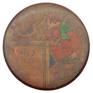 May include: Round, wooden decorative piece with a painted illustration. The image depicts a person in red and green clothing sitting at a table, with a boat-like object on the left. Text is visible in the image. The piece has a dark red border.