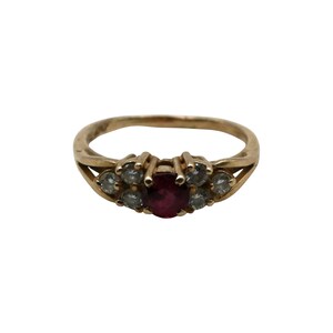 May include: A gold ring featuring a central oval-cut ruby gemstone, flanked by smaller, clear gemstones. The ring has a classic design with a polished finish, suitable for everyday wear or special occasions.