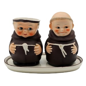May include: A ceramic salt and pepper shaker set featuring two monk figurines in brown robes, each with a smiling face and a white rope belt. The set includes a matching oval tray with a brown rim. The figurines have rosy cheeks and are on a white background.