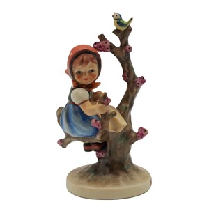 May include: A vintage porcelain figurine of a young girl in a blue dress and red headscarf, seated on a tree branch. A small bird perches on the tree. The figurine has a round base and is painted in soft colors.
