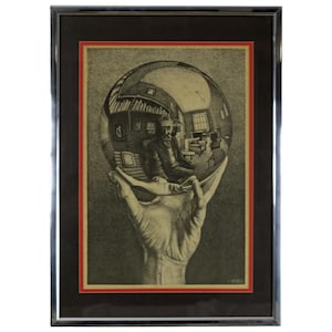 May include: Framed black and white print featuring a hand holding a reflective sphere. Inside the sphere, a man sits in a room with furniture and bookshelves. The print is framed with a black frame and a red border.