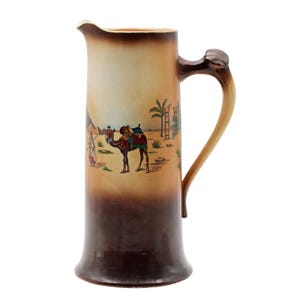 May include: Tall ceramic pitcher with a desert scene featuring a camel, palm trees, and a figure. The pitcher has a brown handle and a gradient color scheme, transitioning from light tan to dark brown.