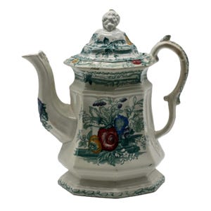 May include: Cream-colored ceramic teapot with a floral design featuring red, blue, and yellow flowers. The teapot has a lid with a decorative knob and a curved handle. Green accents and trim complete the design.