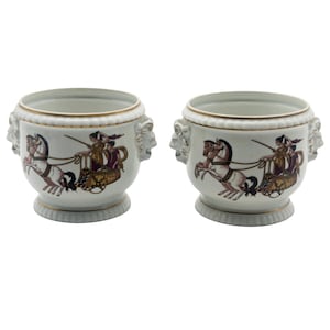 May include: Two white ceramic planters with gold trim and handles shaped like faces. Each planter features a chariot scene with horses and figures in shades of brown, red, and gold. The planters have a pedestal base.