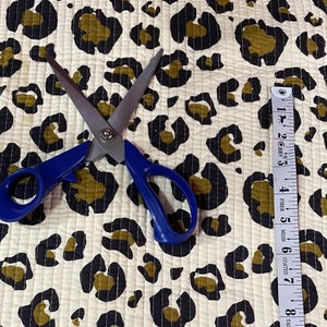 Può includere: Tessuto color crema con stampa leopardata nera e verde oliva. Un paio di forbici con manico blu e un metro a nastro sono sul tessuto. Il metro a nastro mostra misure fino a 35,5 cm.