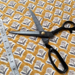 Könnte beinhalten: Geöffnete schwarze Schere mit goldener Niete und weißem Maßband auf gelbem Stoff mit Blumenmuster. Der Stoff hat ein sich wiederholendes Muster aus weißen und grauen Blumenillustrationen.