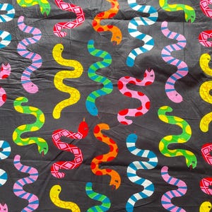 Può includere: Tessuto nero con un motivo ripetuto di serpenti stilizzati colorati. I serpenti sono in vari colori, tra cui rosso, arancione, giallo, verde, blu e rosa, con diversi motivi. I serpenti sono disposti in un motivo ondulato verticale.