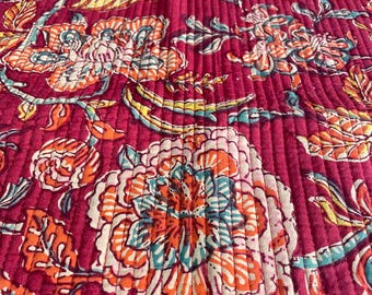 May include: Close-up of a quilted fabric with a floral pattern. The fabric is a deep magenta color with a repeating design of large flowers in shades of orange, teal, and white. The quilting creates a textured, ribbed effect.