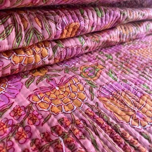 Puede incluir: Primer plano de una pila de tela acolchada rosa con un estampado floral. El diseño presenta flores y hojas naranjas, moradas y verdes. La tela está doblada, mostrando la textura acolchada y el diseño en capas.