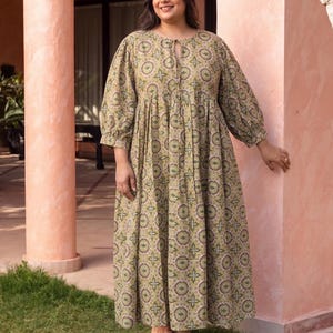 May include: An olive green maxi dress featuring a floral design. The dress has long sleeves and a round neckline. The woman in the image is wearing a white flower in her hair and sandals. The dress is made of a lightweight fabric.