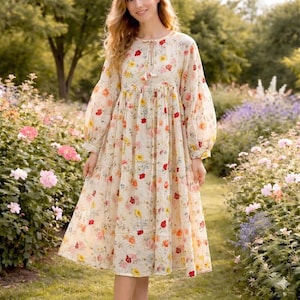 May include: A long-sleeved, cream-colored dress with a floral pattern featuring red, yellow, and pink flowers. The dress has a round neckline with a tie detail and a tiered skirt. The sleeves are long and voluminous.