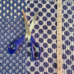 Puede incluir: Un patchwork de tela estampada azul y blanca con unas tijeras azules y una cinta métrica. La tela presenta lunares, círculos y diseños florales. La cinta métrica muestra medidas de hasta 33 cm.