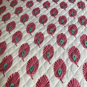 May include: White quilted fabric with a repeating pattern of pink and teal peacock feather designs. The feathers are arranged in rows and columns, creating a symmetrical and visually appealing textile.