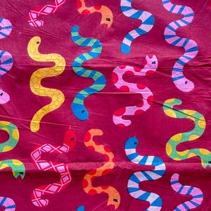 May include: A textile with a repeating pattern of colourful, stylised snakes. The snakes are in shades of yellow, blue, pink, green, and orange, with various patterns, set against a deep red background. The design is playful and eye-catching.