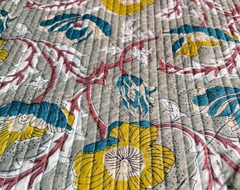 May include: Close-up of a quilted textile with a floral pattern. The design features large yellow and teal flowers with white and red accents on a gray background. The fabric has a stitched, textured appearance.