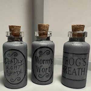 Pode incluir: Três frascos de poção cinzentos com rolhas de cortiça. Cada frasco tem um rótulo com texto preto: "Deadly Night Shade", "Worm's Wort" e "Frog's Breath". Os frascos estão dispostos em fila.