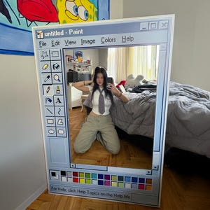 Op de afbeelding: Een grote, rechthoekige spiegel die is ontworpen om op een klassiek Windows Paint-venster te lijken. De spiegelrand is wit, met een blauwe titelbalk en een kleurrijk palet aan de onderkant. De spiegel reflecteert een persoon in een wit overhemd en een kaki broek.