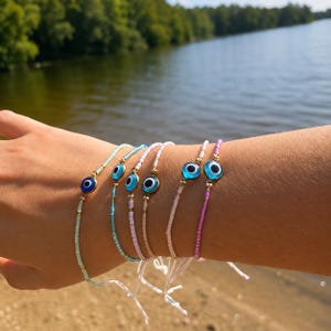 Peut inclure: Un ensemble de cinq bracelets de perles délicats ornés de petits charms en verre en forme d'œil bleu. Les bracelets sont composés de minuscules perles de rocaille aux tons pastel, notamment menthe, bleu clair, rose et blanc, avec des finitions dorées.