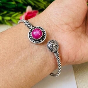 Puede incluir: Un brazalete de tono plateado con un diseño de banda de metal trenzado. Un extremo tiene un engaste circular con una piedra lisa, redonda y de color rosa magenta vibrante, mientras que el otro extremo presenta un disco de metal más pequeño.