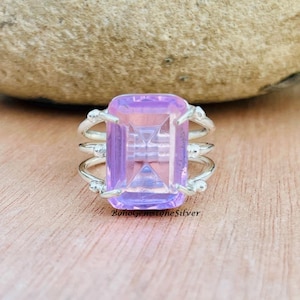 May include: A silver ring with a large, rectangular, light purple gemstone. The gemstone has a geometric design within it. The ring has a triple-band design with small silver accents. The ring is set on a wooden surface.