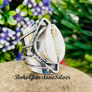 Op de afbeelding: Een zilveren ring met een grote, witte zeeschelp. De ring heeft een brede band met decoratieve zilveren accenten. De tekst "BohoGemstoneSilver" staat onderaan de afbeelding. De ring is geplaatst tegen een natuurlijke achtergrond.
