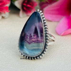 Peut inclure: Une bague en argent ornée d'une grosse pierre en forme de goutte d'eau avec un dégradé de couleurs, dont le bleu, le violet et le blanc. La pierre est sertie dans une lunette en argent avec un bord perlé. Le texte "BohoGems on Silver" est visible.