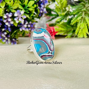 May include: A silver ring with an oval-shaped, multicolored stone. The stone has a pattern of blue, grey, white, and red swirls. The ring is set against a neutral background with the text "BohoGemstoneSilver".