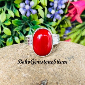 Könnte beinhalten: Ein Silberring mit einem großen, ovalen, leuchtend roten Edelstein. Das Ringband hat ein strukturiertes Design. Der Text "BohoGemstoneSilver" befindet sich am unteren Rand des Bildes. Der Hintergrund zeigt grüne Blätter und bunte Blumen.