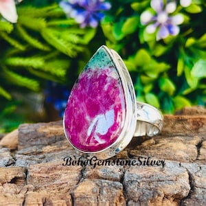 May include: A silver ring with a large, teardrop-shaped gemstone. The gemstone features a vibrant mix of magenta and teal colours. The ring is set on a wooden surface, with the text "BohoGemstoneSilver" visible.