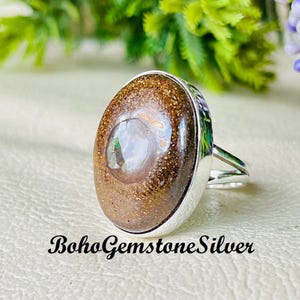 May include: A silver ring featuring a large, oval-shaped gemstone with brown and tan tones. The gemstone has a polished, glossy surface. The ring's band is split at the top, creating a unique design. The text "BohoGemstoneSilver" is at the bottom.