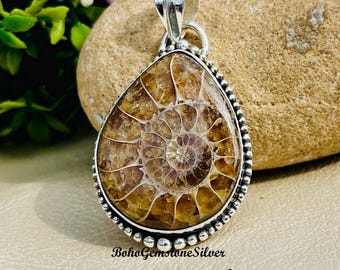 May include: A teardrop-shaped pendant with a fossilized ammonite design. The pendant features a silver-colored bezel with a beaded edge. The fossil is a spiral pattern in shades of brown and tan. The text "BohoGemstoneSilver" is at the bottom.
