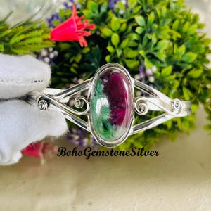以下が含まれることがあります： 緑と赤の色合いが混ざった楕円形の宝石をあしらった、シルバーのカフブレスレット。ブレスレットはスクロールワークの装飾が施され、「BohoGemstoneSilver」の文字が入っています。
