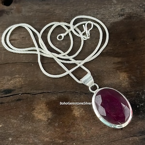 May include: A silver necklace featuring a large, oval, faceted ruby pendant. The ruby is a deep red hue, set in a silver bezel. The chain is a delicate, woven design. The text "BohoGemstoneSilver" is visible.