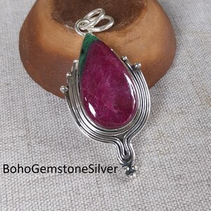 Puede incluir: Colgante de plata con una gran piedra preciosa de rubí en forma de lágrima. El rubí es de color magenta intenso con un acento verde en la parte superior. El colgante tiene intrincados detalles plateados y un lazo para una cadena. El texto "BohoGemstoneSilver" es visible.