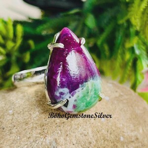 May include: A silver ring featuring a large, teardrop-shaped gemstone. The gemstone displays a blend of colors, including deep purple, green, and white. The ring is set on a neutral-toned surface, with the text "BohoGemstoneSilver" visible.