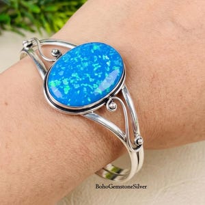 Puede incluir: Un brazalete de plata con una gran piedra de ópalo azul de forma ovalada con un patrón moteado vibrante. La banda de metal tiene un acabado pulido y está diseñada con delicados detalles de alambre en espiral que enmarcan la piedra.