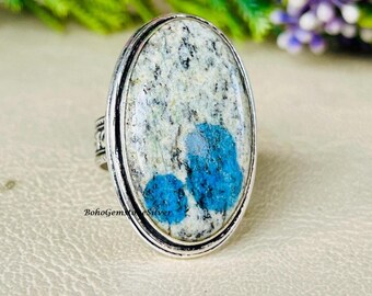 May include: Oval silver ring featuring a large, light-colored stone with blue and gray patterns. The stone has two prominent blue sections. The ring's band has decorative detailing. The text "BohoGemsSilver" is visible on the ring.