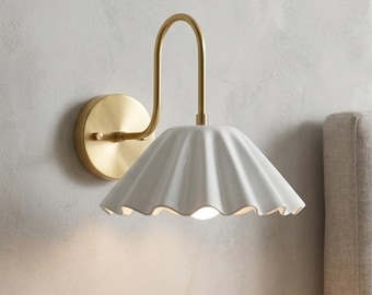 May include: A wall-mounted lamp featuring a brushed brass finish arm and a circular wall plate. The lamp has a white, fluted, bell-shaped shade with a scalloped edge. The light fixture is positioned above a small dark vase and a brown leather notebook.