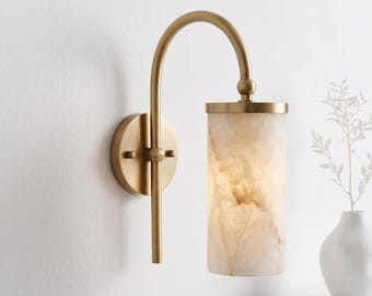 May include: A wall-mounted light fixture featuring a brushed brass finish metal arm with a curved design and a cylindrical shade made of translucent, cream-colored stone with natural veining patterns that emit a soft, warm glow.