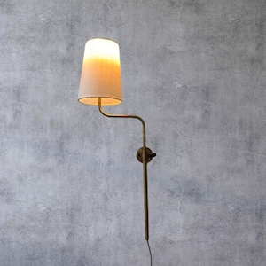May include: A wall-mounted lamp with a beige lampshade and a brass-colored arm and base. The lampshade is a light beige color and the light is on. The lamp is mounted on a gray wall.