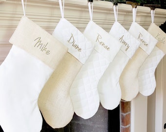 May include: Six white and beige Christmas stockings with names embroidered on them: Mike, Diane, Renae, Avery, Reese, and Koda.