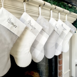 Puede incluir: Seis calcetines navideños personalizados en tonos grises y blancos cuelgan de una repisa. Cada calcetín tiene un nombre bordado en el puño: Homer, Marge, Bart, Lisa, Maggie y Snowball.