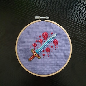 以下が含まれることがあります： 木製のフレームに入ったクロスステッチ刺繍。青い刃と赤と金の柄を持つピクセル化された剣が特徴で、ラベンダー色の背景に赤いピクセル化された血しぶきが散らばっています。フレームは金属製の留め金で固定されています。