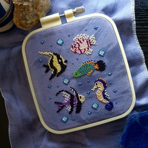 May include: A square embroidery hoop containing a light purple fabric with a cross-stitch design of four colourful fish and a seahorse surrounded by small blue bubbles. The hoop has a white plastic frame with a metal tightening screw at the top.