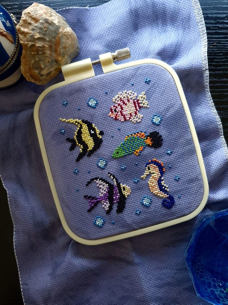 May include: A square embroidery hoop containing a light purple fabric with a cross-stitch design of four colourful fish and a seahorse surrounded by small blue bubbles. The hoop has a white plastic frame with a metal tightening screw at the top.