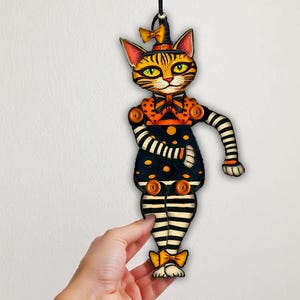以下が含まれることがあります： 黒とオレンジの水玉模様とストライプ柄の関節付き猫のオーナメント。猫は魔女の帽子と蝶ネクタイを身に着けており、関節部分にはオレンジ色のボタンが付いています。Printable Halloween Articulated Catsというテキストが表示されています。