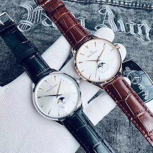 May include: Two luxury wristwatches with leather bands. One watch has a silver face and a black band, the other has a white face and a brown band. Both watches have a moon phase indicator and gold-colored hands.