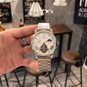 May include: A silver wristwatch with a white face, Roman numerals, and a visible mechanical movement. The watch has a silver band and is held by a hand. The watch face includes smaller dials and a moon phase indicator. The watch is a luxury item.